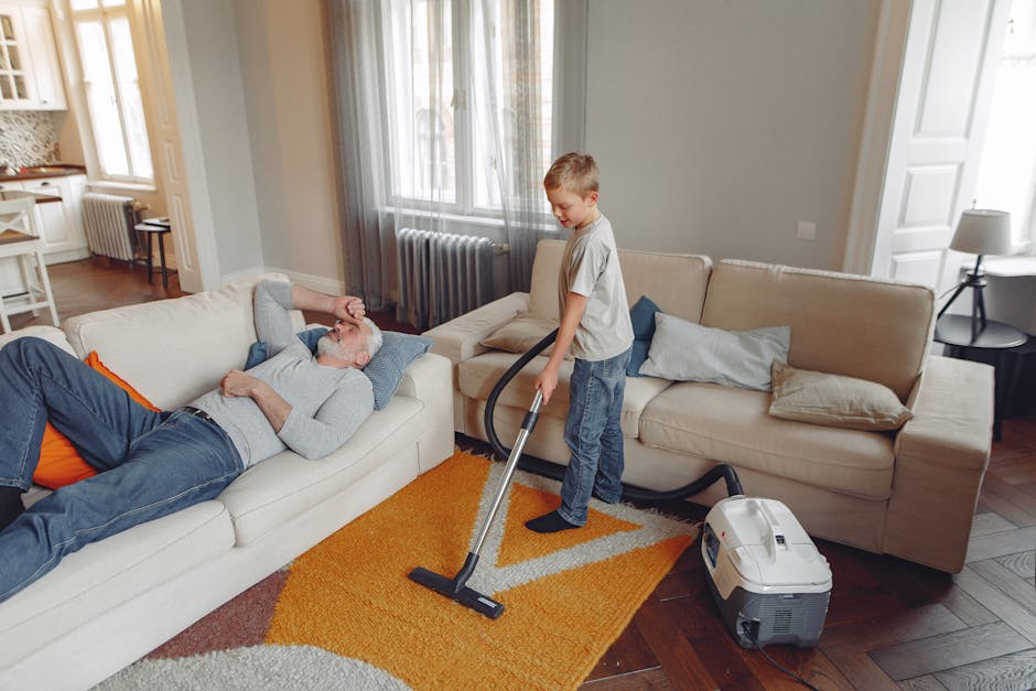 In a well-lit living room with large windows, a young boy is vacuuming a vibrant orange and beige rug placed on a dark wooden floor. He is holding a modern, white vacuum cleaner with a grey hose, whil