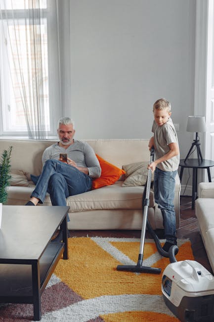 In a well-lit living room with large windows, a young boy is vacuuming a vibrant orange and beige rug placed on a dark wooden floor. He is holding a modern, white vacuum cleaner with a grey hose, whil