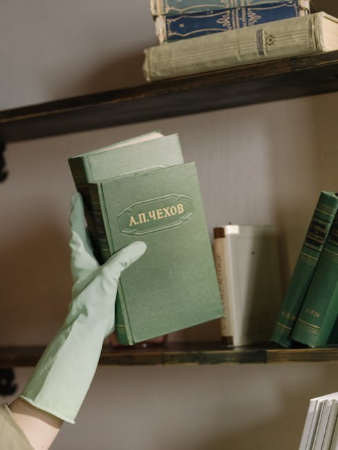 A woman with curly hair tied back with a patterned headscarf is inspecting or arranging white booklets or magazines. She is dressed in a light green shirt and wearing pale green gloves, standing in a room with a dark wooden ceiling and shelving unit. The shelves hold framed artwork, vases, and stacked books. The background features a brick wall and soft natural lighting, emphasizing a clean and organized space suitable for domestic or commercial cleaning reflection. The setting highlights attention to detail and maintenance of a tidy environment, aligning with surface cleaning and deep cleaning practices suggested by the webpage title.