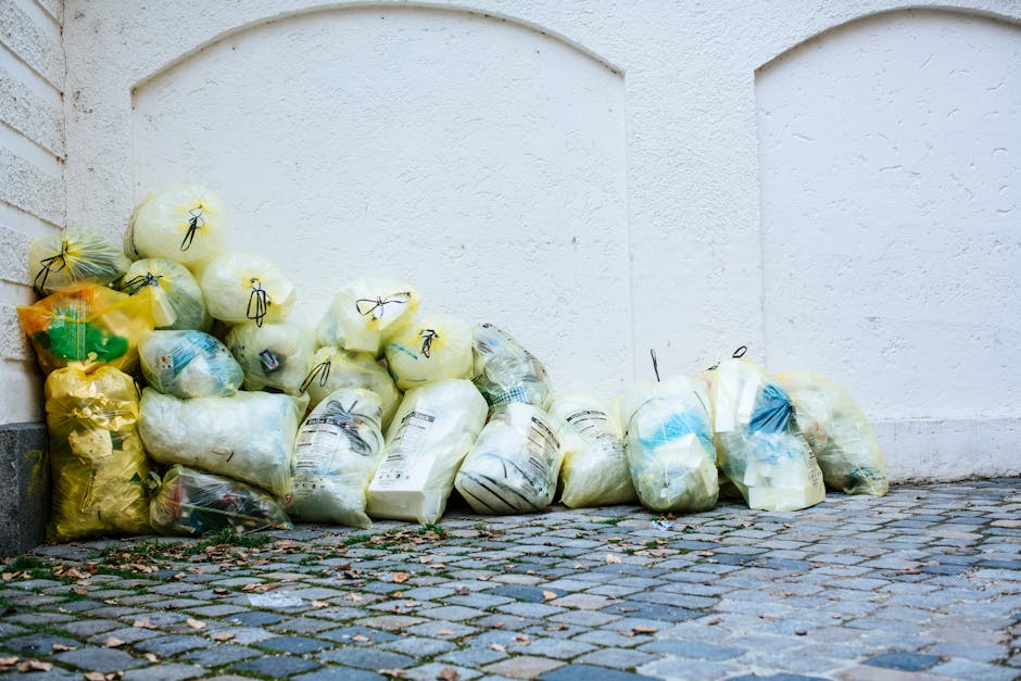 A row of yellow and white plastic bags filled with waste, placed on a cobblestone pavement next to a white-painted exterior wall of a building. The bags contain various types of rubbish and are tied c