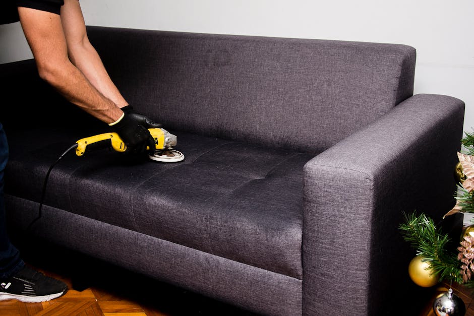 A woman with dark hair, wearing a black uniform, stands in a well-lit living room holding beige cushions, indicating she is preparing to clean or refresh upholstery. The room features a white sofa wit